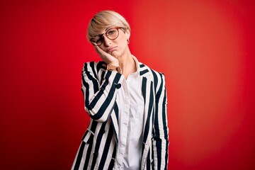 Blonde business woman with short hair wearing glasses and striped jacket over red background thinking looking tired and bored with depression problems with crossed arms.