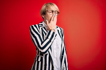 Blonde business woman with short hair wearing glasses and striped jacket over red background bored yawning tired covering mouth with hand. Restless and sleepiness.