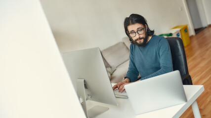 Multitasking. Young caucasian concentrated bearded man sitting at his workplace and working remotely, using desktop computer and laptop