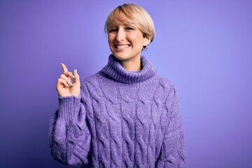 Young blonde woman with short hair wearing winter turtleneck sweater over purple background with a big smile on face, pointing with hand finger to the side looking at the camera.