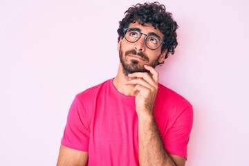 Handsome young man with curly hair and bear wearing casual clothes and glasses serious face thinking about question with hand on chin, thoughtful about confusing idea