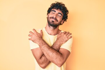 Handsome young man with curly hair and bear wearing casual tshirt over yellow background hugging...