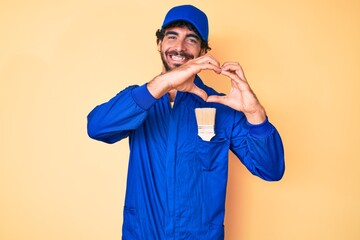 Handsome young man with curly hair and bear wearing builder jumpsuit uniform smiling in love doing heart symbol shape with hands. romantic concept.