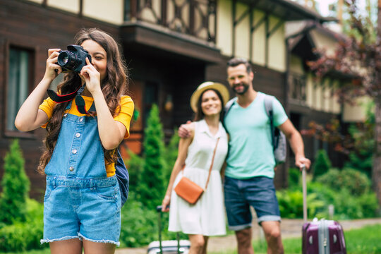 Nice photo. A photo of a happy travelling family with suitcases with a hotel in their background, blurred parents hugging, daughter in overalls staying in the foreground, making a photo.