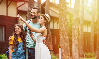 Summer journey. A photo with sunny light effect of a happy family on a weekend staying near hotel, smiling daughter and father looking the way mother pointing with her hand.