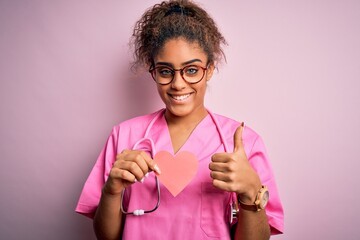 African american cardiologist girl wearing medical uniform and stethoscope holding heart happy with big smile doing ok sign, thumb up with fingers, excellent sign