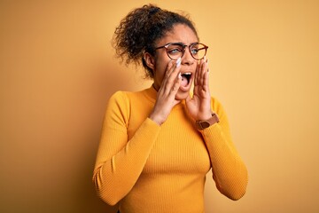 Young beautiful african american girl wearing sweater and glasses over yellow background Shouting angry out loud with hands over mouth