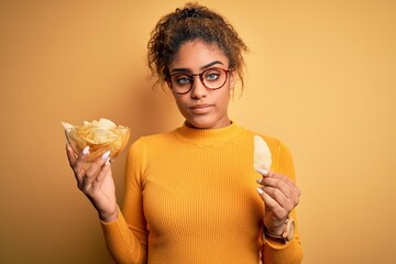 Young african american afro girl holding bowl with potatoes chips over yellow background with a...