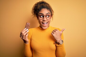 Young african american afro girl eating energetic protein bar of cereal over yellow background...