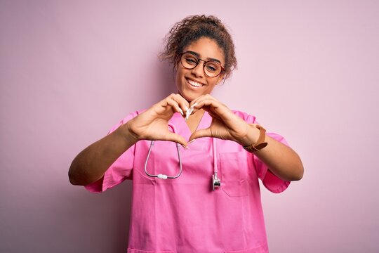 African american nurse girl wearing medical uniform and stethoscope over pink background smiling in love doing heart symbol shape with hands. Romantic concept.