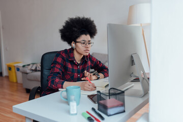 Side view of young focused afro american female student looking at computer screen and making some notes while learning online at home