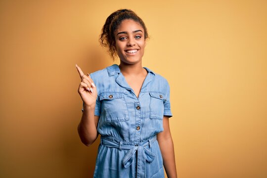 Young beautiful african american girl wearing denim dress standing over yellow background with a big smile on face, pointing with hand finger to the side looking at the camera.