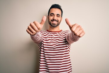 Fototapeta premium Young handsome man with beard wearing casual striped t-shirt standing over white background approving doing positive gesture with hand, thumbs up smiling and happy for success. Winner gesture.