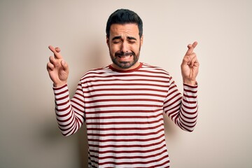 Young handsome man with beard wearing casual striped t-shirt standing over white background gesturing finger crossed smiling with hope and eyes closed. Luck and superstitious concept.