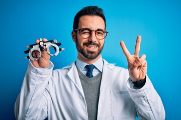 Young handsome optical man with beard holding optometry glasses over blue background smiling with happy face winking at the camera doing victory sign with fingers. Number two.