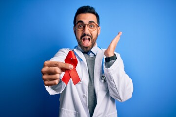 Young handsome doctor man with beard wearing stethoscope holding red hiv ribbon very happy and excited, winner expression celebrating victory screaming with big smile and raised hands