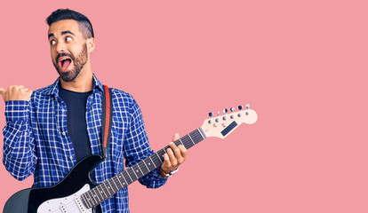 Young hispanic man playing electric guitar pointing thumb up to the side smiling happy with open mouth