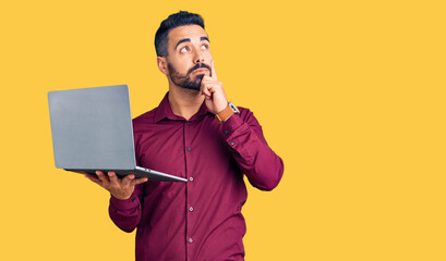 Young hispanic man holding laptop serious face thinking about question with hand on chin,...