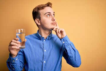 Young handsome redhead man drinking glass of water over isolated yellow background serious face...
