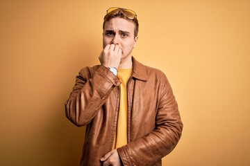 Young handsome redhead man wearing casual leather jacket over isolated yellow background looking stressed and nervous with hands on mouth biting nails. Anxiety problem.