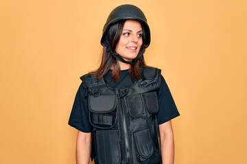 Young beautiful brunette soldier woman wearing bulletproof and security helmet looking away to side with smile on face, natural expression. Laughing confident.