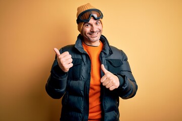 Young handsome skier man skiing wearing snow sportswear using ski goggles Pointing to the back behind with hand and thumbs up, smiling confident