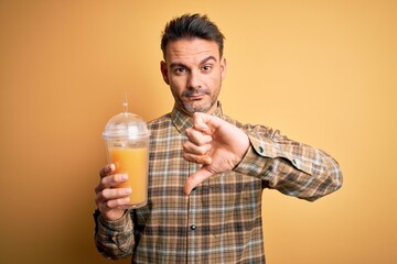 Young handsome man drinking healthy orange juice using straw over yellow background with angry face, negative sign showing dislike with thumbs down, rejection concept