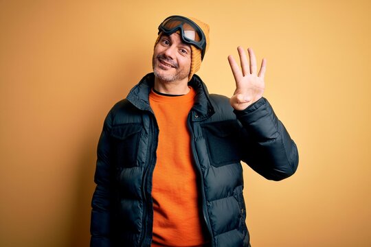 Young handsome skier man skiing wearing snow sportswear using ski goggles showing and pointing up with fingers number four while smiling confident and happy.