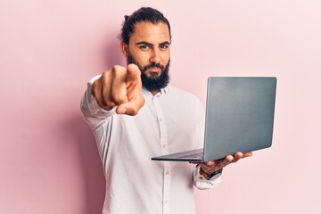 Young arab man holding laptop pointing with finger to the camera and to you, confident gesture...