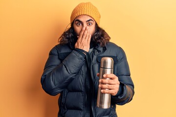 Young arab man holding thermo covering mouth with hand, shocked and afraid for mistake. surprised...