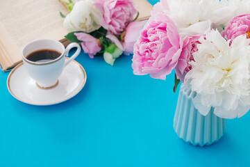 Summer romantic breakfast. Blue vase with peonies, white cup of coffee, open book on a blue background.Good morning concept.Copy space, selective focus with shallow depth of field