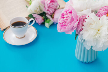 Summer romantic breakfast. Blue vase with peonies, white cup of coffee, open book on a blue...
