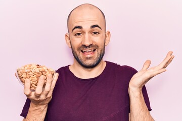 Young handsome bald man holding bowl with peanuts celebrating achievement with happy smile and winner expression with raised hand