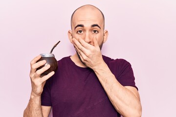 Young handsome bald man drinking mate infusion beverage covering mouth with hand, shocked and...