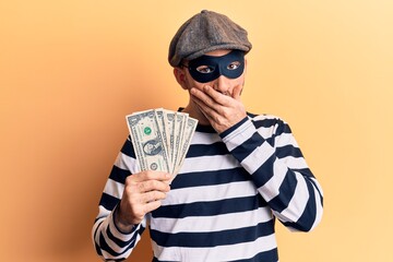 Young handsome bald man wearing burglar mask stealing dollars banknotes covering mouth with hand, shocked and afraid for mistake. surprised expression