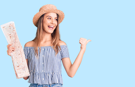 Young beautiful girl wearing hat holding city map pointing thumb up to the side smiling happy with open mouth