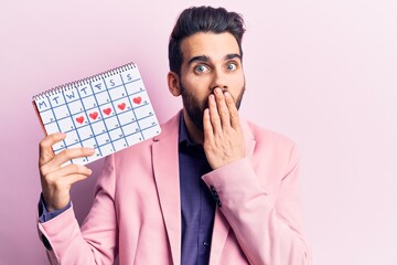 Young handsome man with beard holding calendar with hearts covering mouth with hand, shocked and...