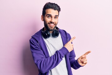 Young handsome man with beard listening to music using headphones smiling and looking at the camera pointing with two hands and fingers to the side.