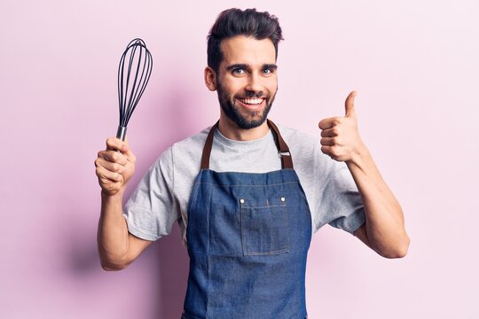 Young handsome man with beard wearing apron holding whisk smiling happy and positive, thumb up doing excellent and approval sign