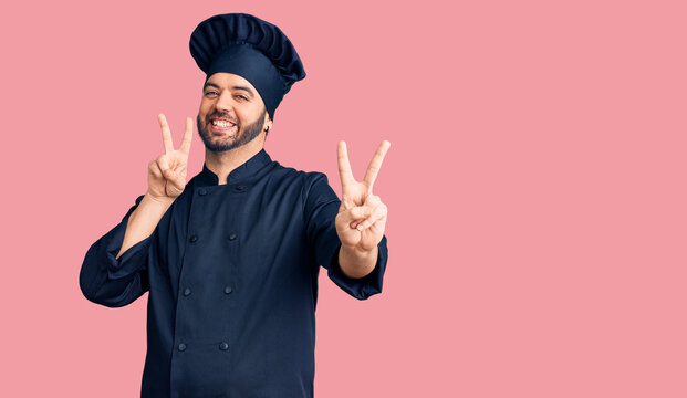 Young hispanic man wearing cooker uniform smiling looking to the camera showing fingers doing victory sign. number two.