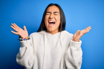 Young beautiful asian sportswoman wearing sweatshirt standing over isolated blue background celebrating mad and crazy for success with arms raised and closed eyes screaming excited. Winner concept