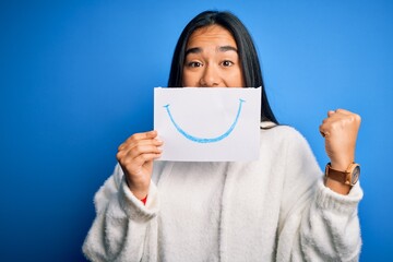 Young beautiful asian woman holding paper with smile draw on mouth over blue background screaming proud and celebrating victory and success very excited, cheering emotion