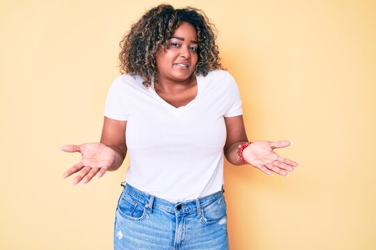 Young african american plus size woman wearing casual white tshirt clueless and confused expression with arms and hands raised. doubt concept.