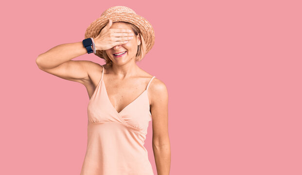 Young blonde woman wearing summer hat smiling and laughing with hand on face covering eyes for surprise. blind concept.