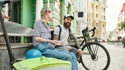 Two cheerful couriers, young man and woman sitting on the bench and talking outdoors while...