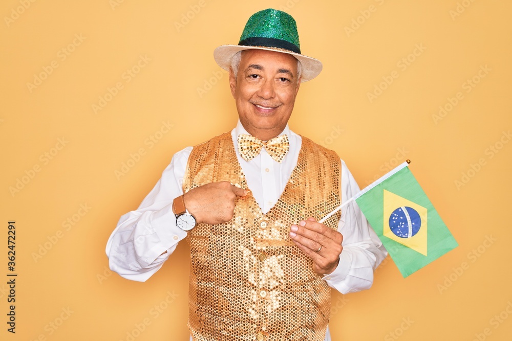 Wall mural Middle age senior grey-haired man wearing Brazilian carnival custome holding Brazil flag with surprise face pointing finger to himself