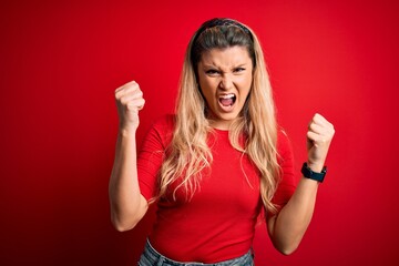 Young beautiful blonde woman wearing casual t-shirt standing over isolated red background angry and...