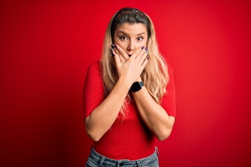 Young beautiful blonde woman wearing casual t-shirt standing over isolated red background shocked covering mouth with hands for mistake. Secret concept.
