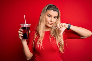 Young beautiful blonde woman drinking cola fizzy beverage using straw over red background with...
