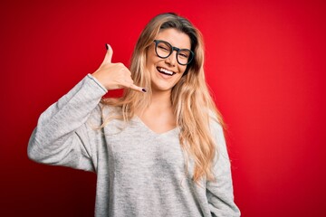 Young beautiful blonde woman wearing sweater and glasses over isolated red background smiling doing phone gesture with hand and fingers like talking on the telephone. Communicating concepts.